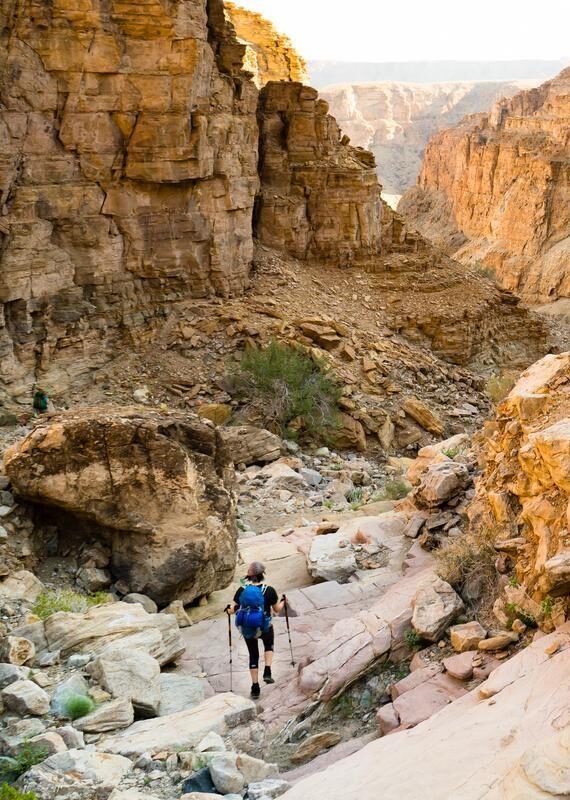 Person with hiking gear walking through a rugged, boulder-strewn canyon floor between high orange cliffs.