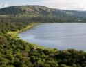 Wide view of a blue lake in Akagera National Park with lush green trees, safari tents, and hills under a clear sky.