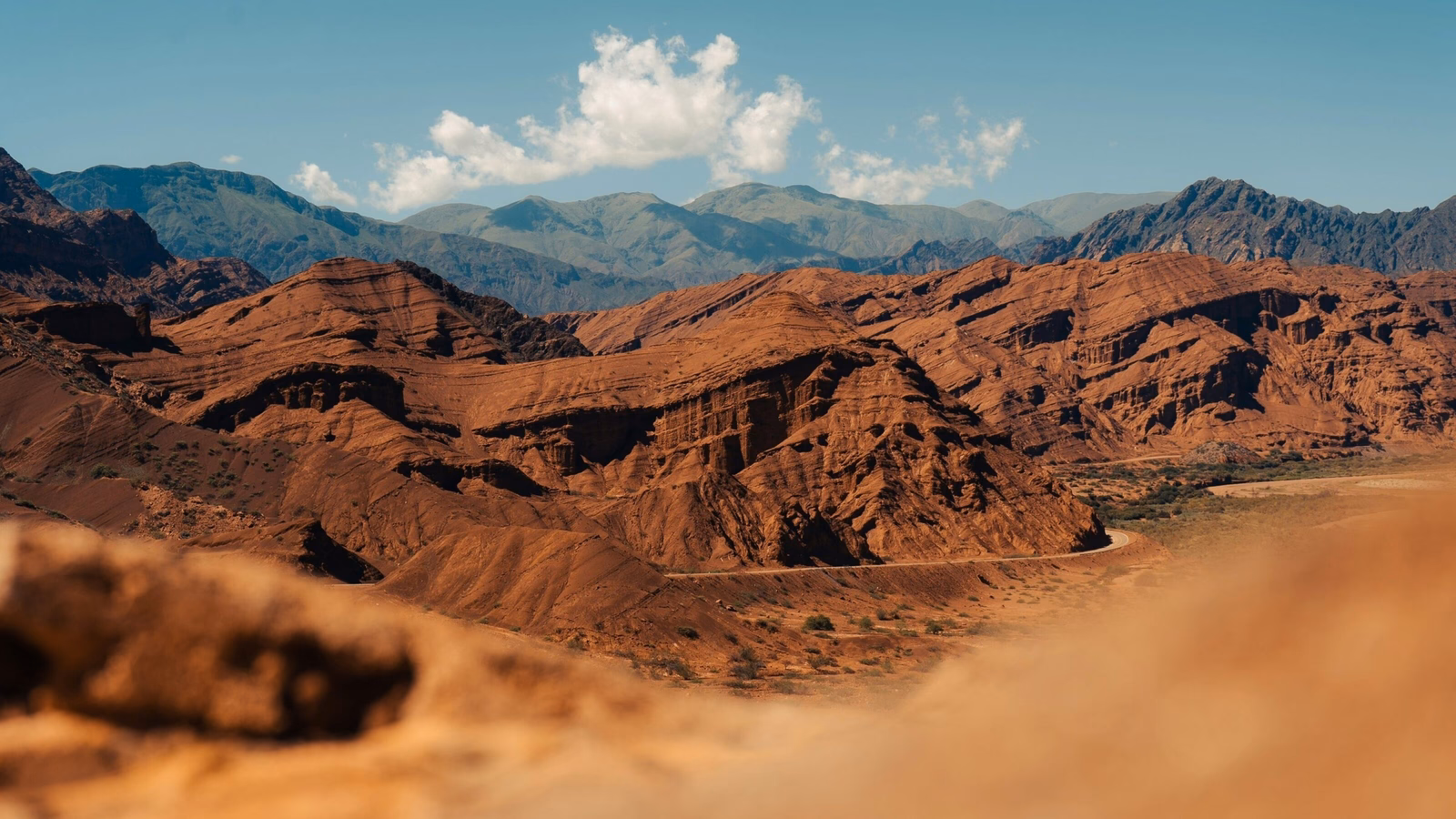 Jagged red rock mountains and desert terrain under a clear sky with distant blue mountain ranges in Salta, Argentina.