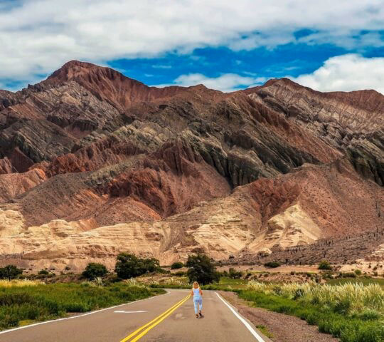 A paved road leads toward layered red and purple mountains with a person walking in the distance in Salta, Argentina.