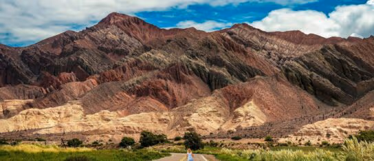 A paved road leads toward layered red and purple mountains with a person walking in the distance in Salta, Argentina.