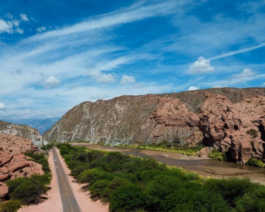 Aerial view of a paved road running through a red rock canyon and green valley under a bright blue sky.