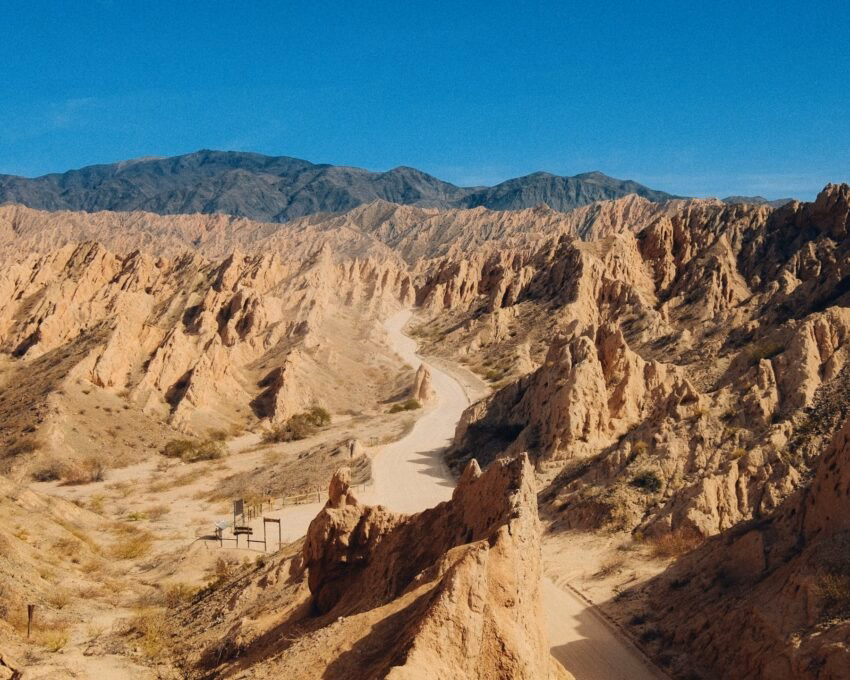 Narrow dirt road winding through sharp, angled sandstone rock formations in a desert canyon under a clear sky.