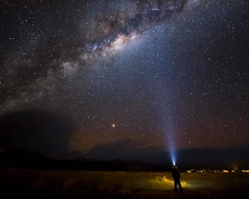 Silhouette of a person shining a light beam into a brilliant, star-filled night sky featuring the Milky Way galaxy.