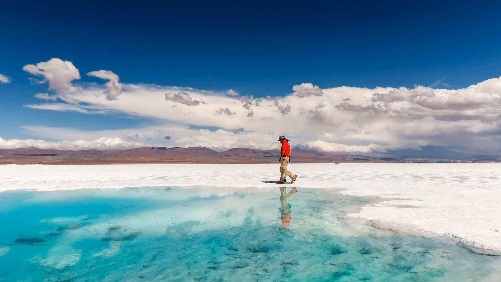 Person walking near a bright turquoise water pool on white salt flats with mountain reflections under a blue sky.