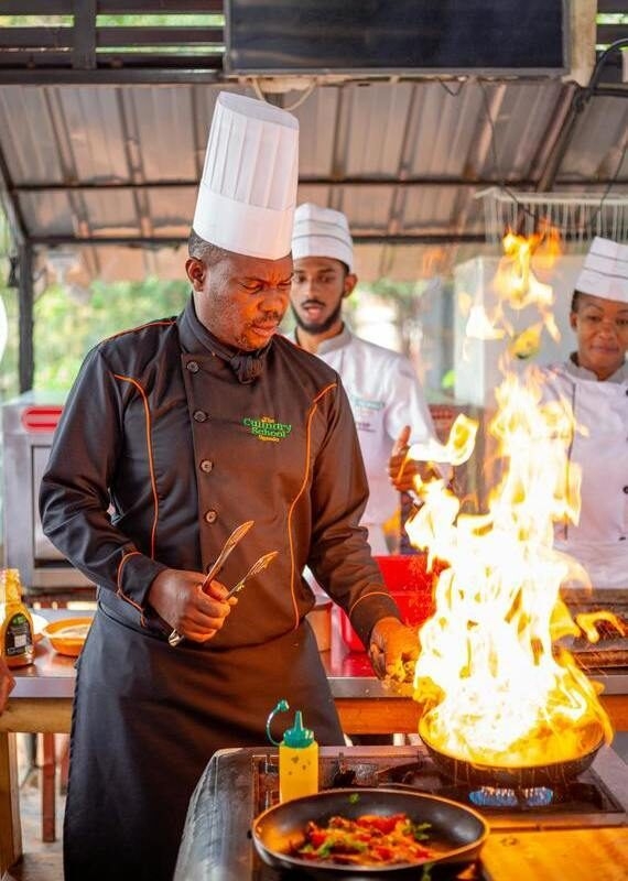 A chef at The Culinary School Uganda prepares a meal with a large burst of fire in a frying pan.