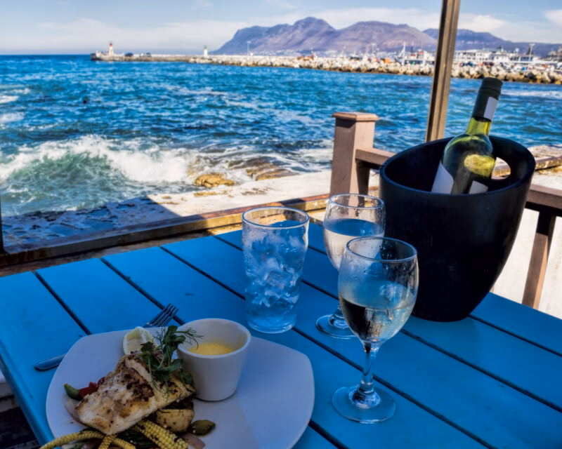 Grilled fish dish and white wine on a blue wooden table at a seaside restaurant with ocean waves in the background.