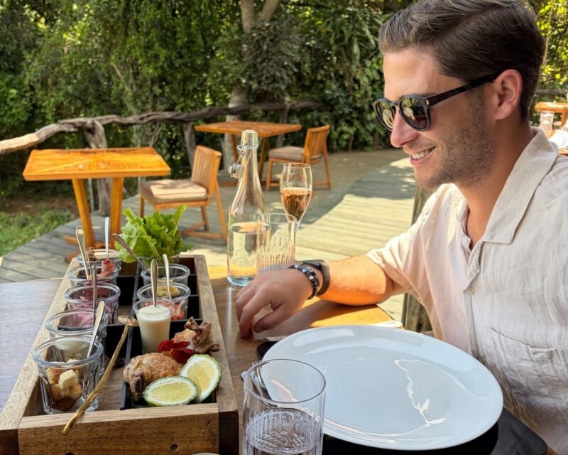 A man points to a wooden tasting tray filled with small jars of food, served with lime and drinks on an outdoor deck.