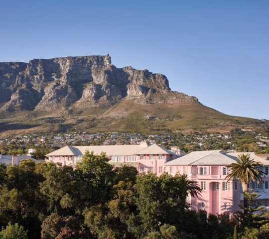 Exterior view of a grand pink hotel surrounded by trees with the flat-topped Table Mountain in the distance.