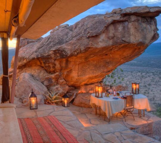 A candlelit dinner table set on a rocky terrace under a giant boulder at Saruni Samburu honeymoon villa.