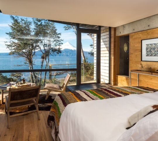 Modern hotel bedroom with a large window view of a blue lake and a snow-capped volcano under a clear sky.