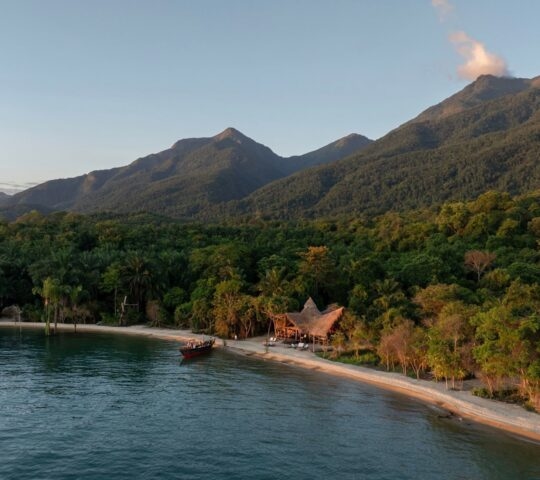 Aerial view of a remote beach lodge with thatched roofs on a white sand beach surrounded by dense jungle and mountains.