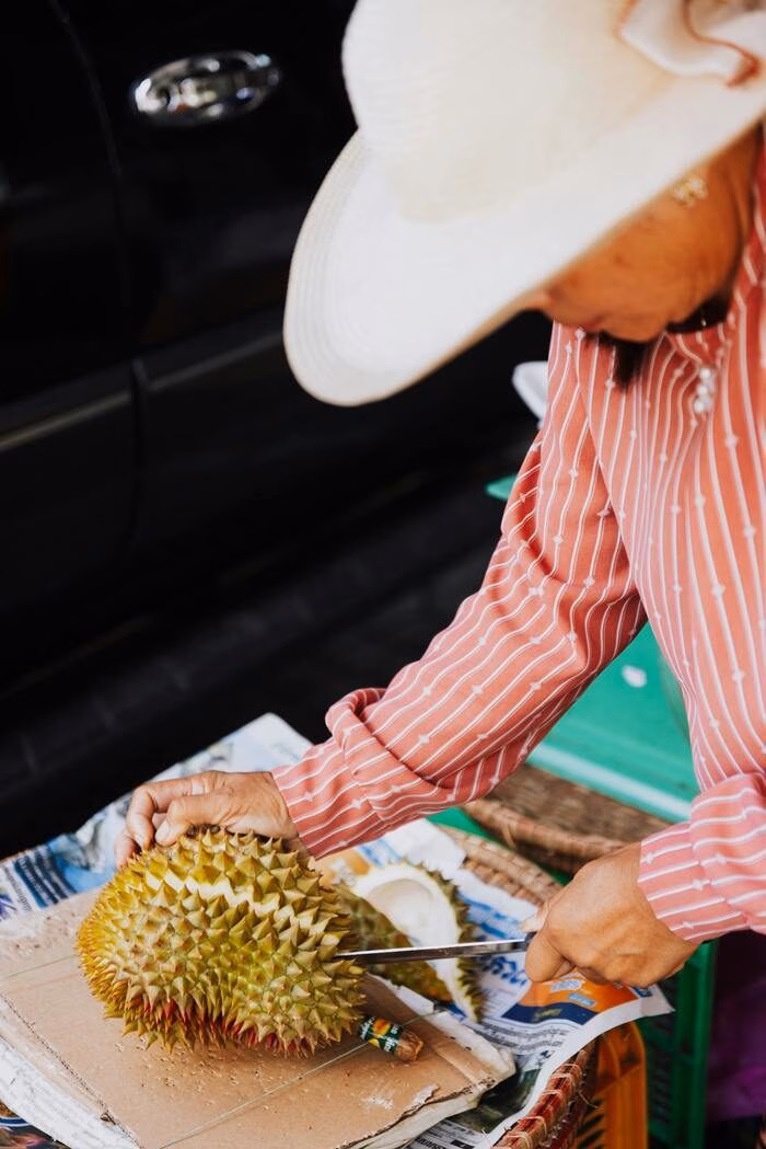 Close-up of a person slicing into a large, green spiky durian fruit at an outdoor market.