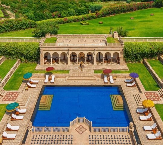 Symmetrical swimming pool at The Oberoi Amarvilas surrounded by green lawns, stone terraces, and white lounge chairs.