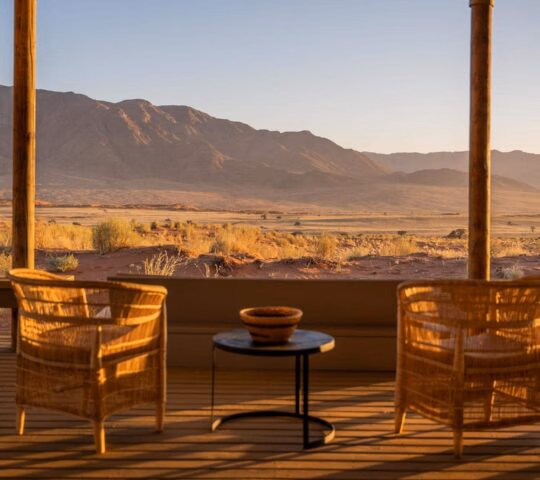 Wicker chairs on a wooden terrace overlooking a desert valley and mountains at sunset.