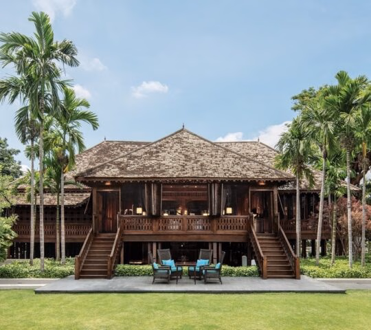 A large, traditional dark wood Thai building with twin staircases and a symmetrical facade surrounded by palm trees.