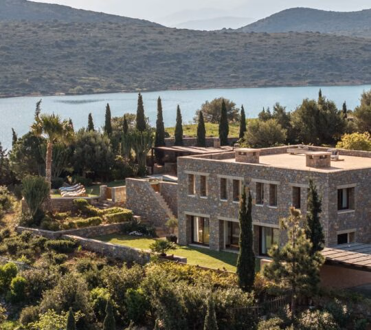 Aerial view of a villa at Phaea Blue Palace in Crete with surrounding trees and water in the background