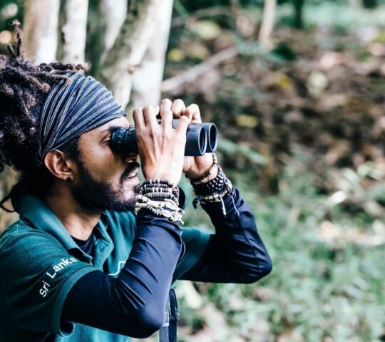 A man with dreadlocks and a headband looking through binoculars in a wooded area.