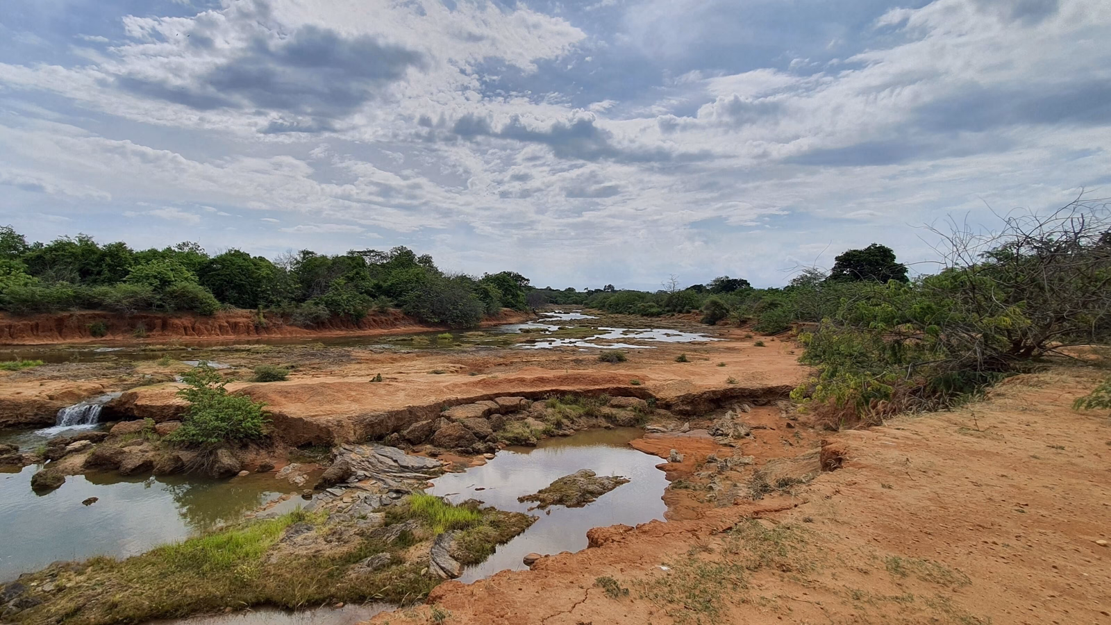 A rocky riverbed with small pools of water surrounded by red soil and green bushes under a vast sky.