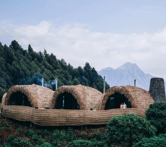 Cocoon-shaped villas at Bisate Reserve sit on a forested hillside overlooking a distant mountain peak under a cloudy sky.