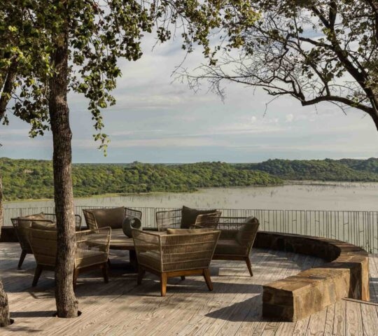 An outdoor patio with comfortable seating overlooking a large lake surrounded by dense green forest and hills.