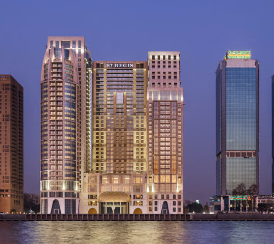 The St. Regis Cairo hotel and neighboring skyscrapers reflecting in the Nile River at dusk under a deep blue sky.
