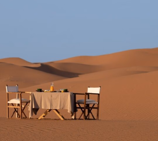 A breakfast table and two chairs set in the middle of vast desert sand dunes under a clear blue sky.