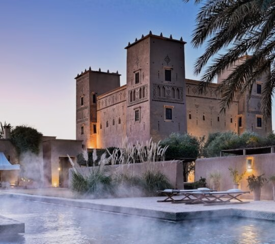 A steaming swimming pool at dusk with a large, lit-up Moroccan kasbah and palm trees in the background.