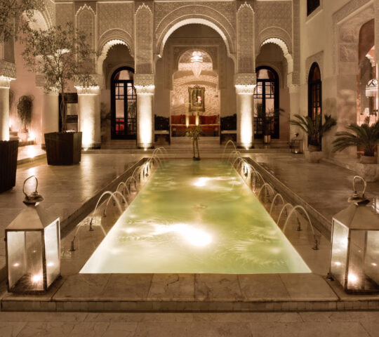 An ornate Moroccan courtyard at night with a glowing narrow pool, water fountains, and arched architecture.