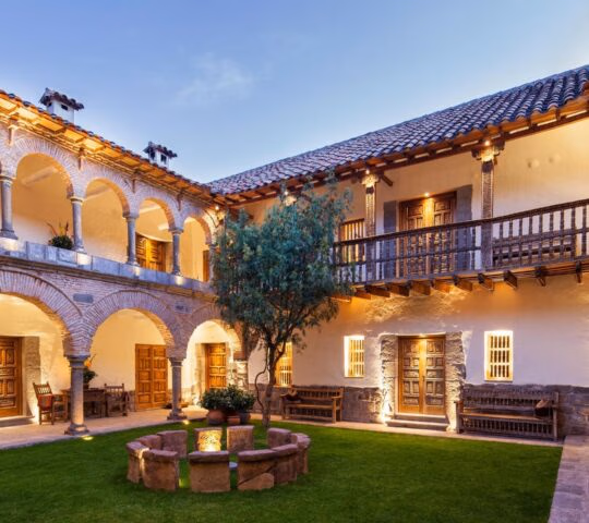 An illuminated colonial courtyard with stone archways, a green lawn, and wooden balconies at Inkaterra La Casona.