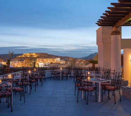 An outdoor restaurant terrace with set tables and candles overlooking a lit-up hillside town at dusk.