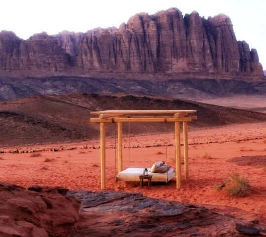 A wooden four-poster daybed on red desert sand with massive rock formations in the background at Discovery Bedu.