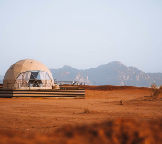 A geodesic dome tent at Sun City Camp in Wadi Rum, Jordan, nestled against jagged desert rock formations under bright sun.