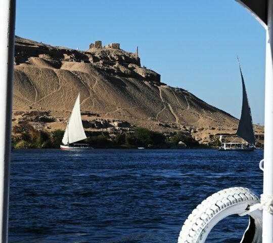 View from a boat of two felucca sailboats on the Nile River with desert hills in the background.