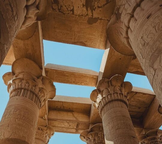 Upward view of ancient Egyptian temple columns with floral capitals and hieroglyphs under a bright sky.