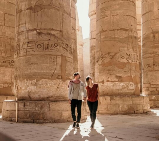 A man and woman walk through a forest of massive ancient Egyptian stone columns covered in carvings.