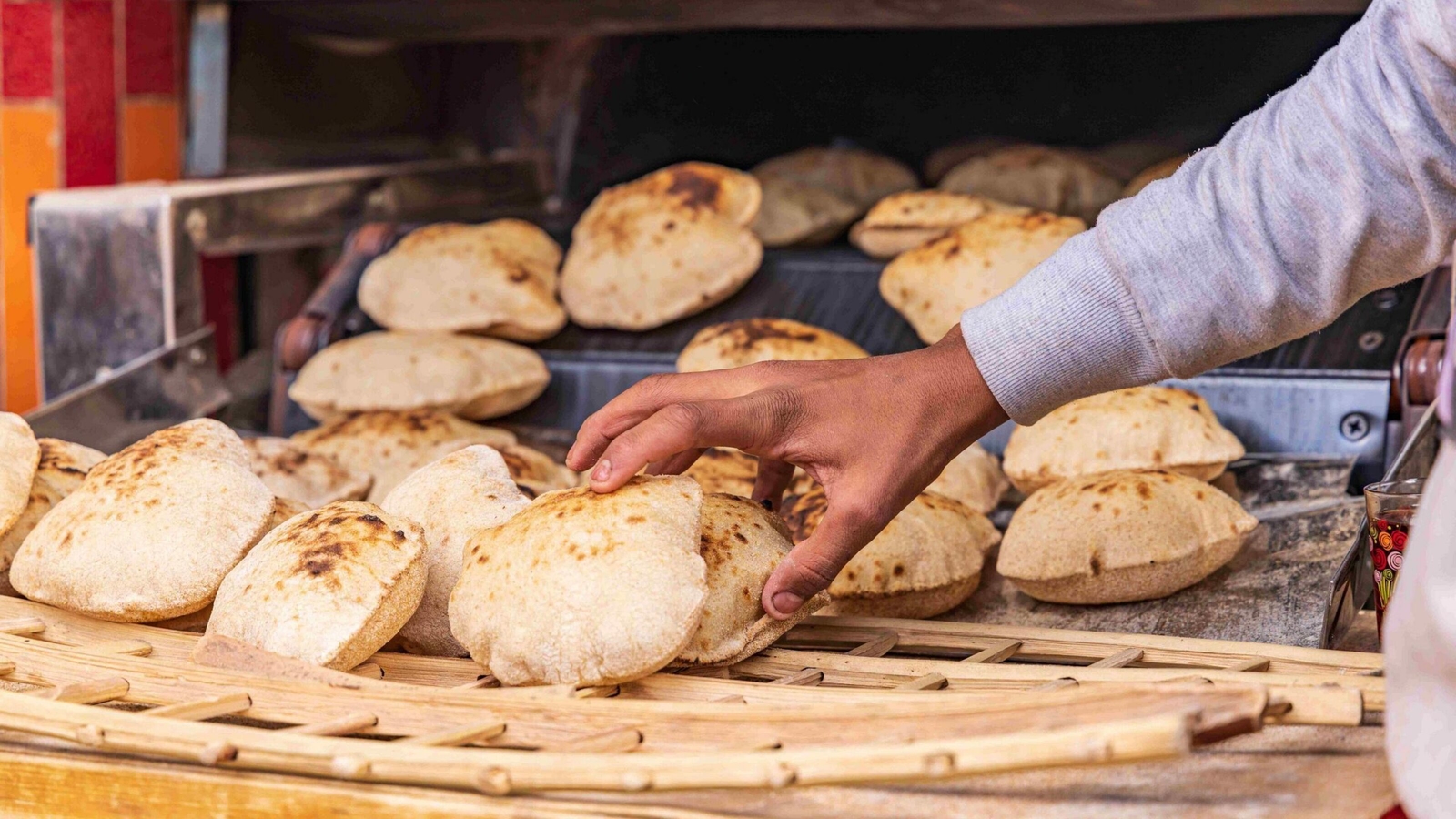 A hand picks up a piece of warm, round flatbread from a wooden rack at an Egyptian bakery.