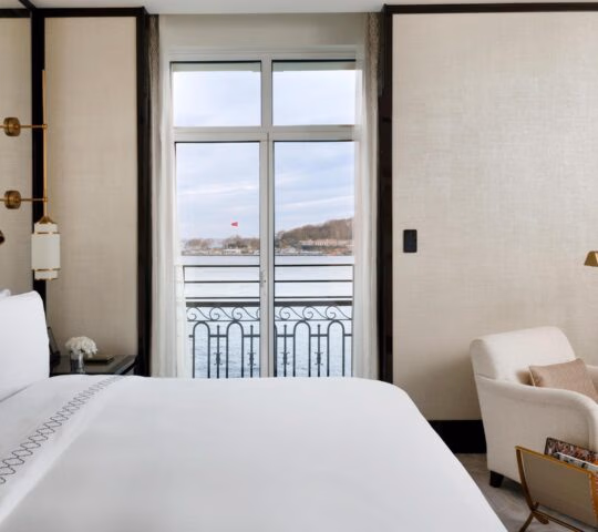 A modern white hotel bedroom with an armchair and glass doors opening to a view of the water and a distant flag.