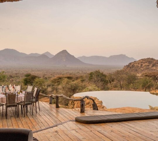 A dining table with chairs on a wooden deck facing a swimming pool and distant mountains under a dusky sky.