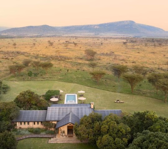 High-angle view of Amani House surrounded by trees overlooking a wide, grassy plain and distant mountains.