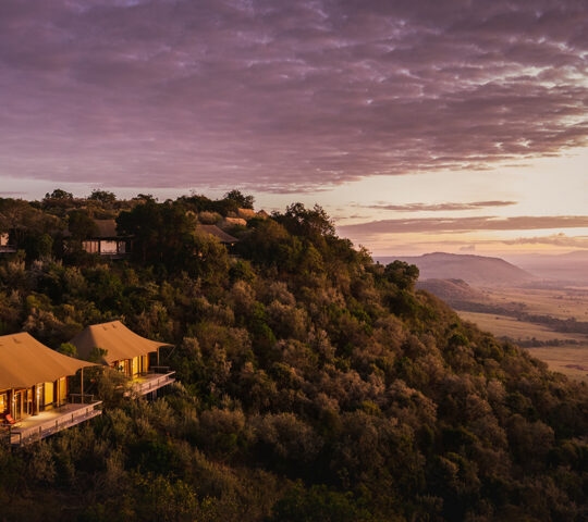 Luxury safari tents on a green hillside at sunset overlooking a wide African savannah landscape.