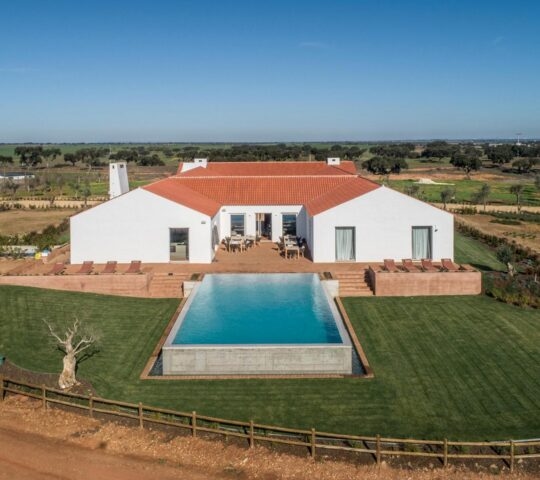 Aerial view of a white villa with terracotta roff and outdoor swimming pool surrounded by green fields at Herdade da Malhadinha Nova