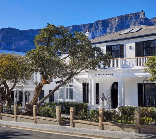 A white two-story boutique hotel with black shutters, captioned Cape Cadogan, with Table Mountain in the background.