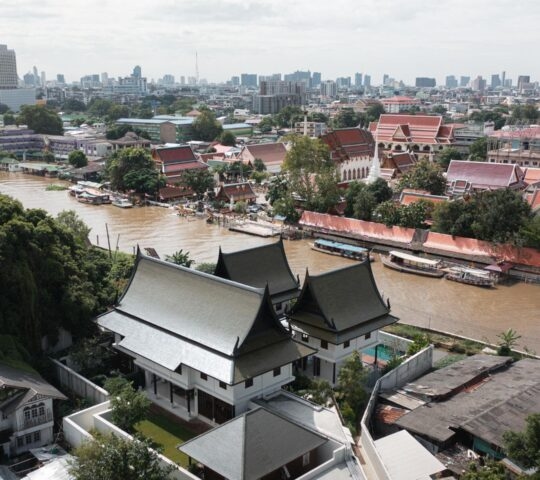 Aerial view of a wide canal in Bangkok with traditional Thai temples, lush trees, and city skyline in the distance.