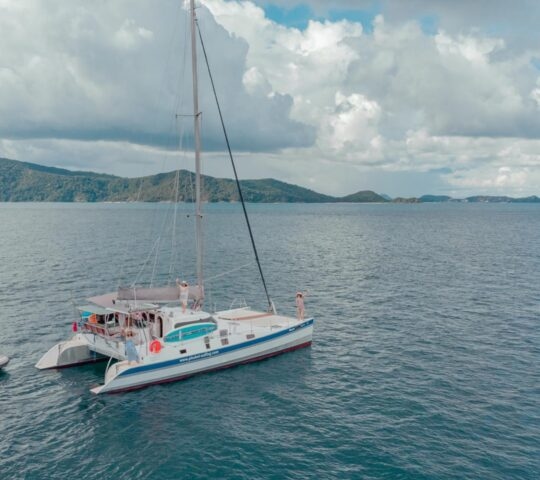 A white catamaran yacht with passengers on deck sailing on blue water with islands and clouds in the background.