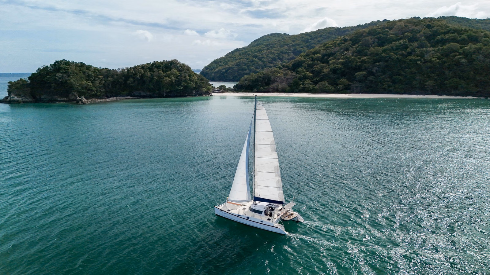 A white catamaran with sails up navigating clear green water near a tropical island with a white sand beach.