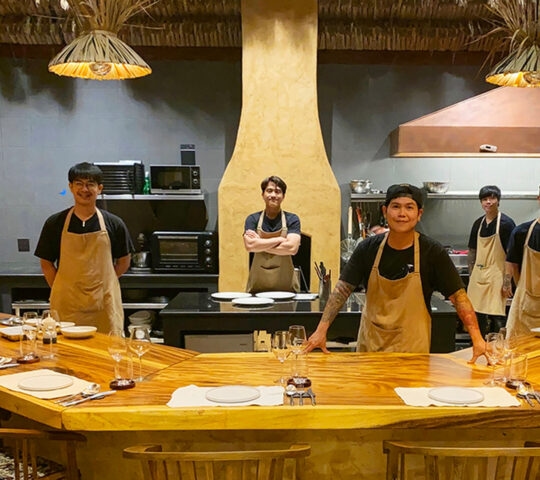 Five chefs stand behind a long, U-shaped wooden table set with plates and wine glasses in a rustic, modern kitchen.