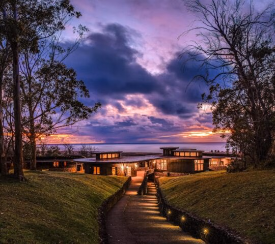 A lighted modern hotel building at dusk with a stone path leading toward it under a vibrant purple and orange sunset sky.