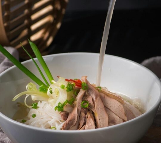 Hot broth being poured into a bowl of Vietnamese noodle soup with meat, spring onions, and sprouts.