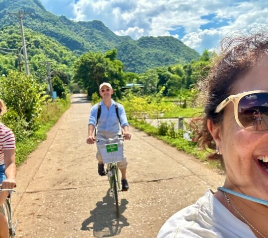 A group of three people smiling and laughing while riding bikes on a sunny path through lush green hills.